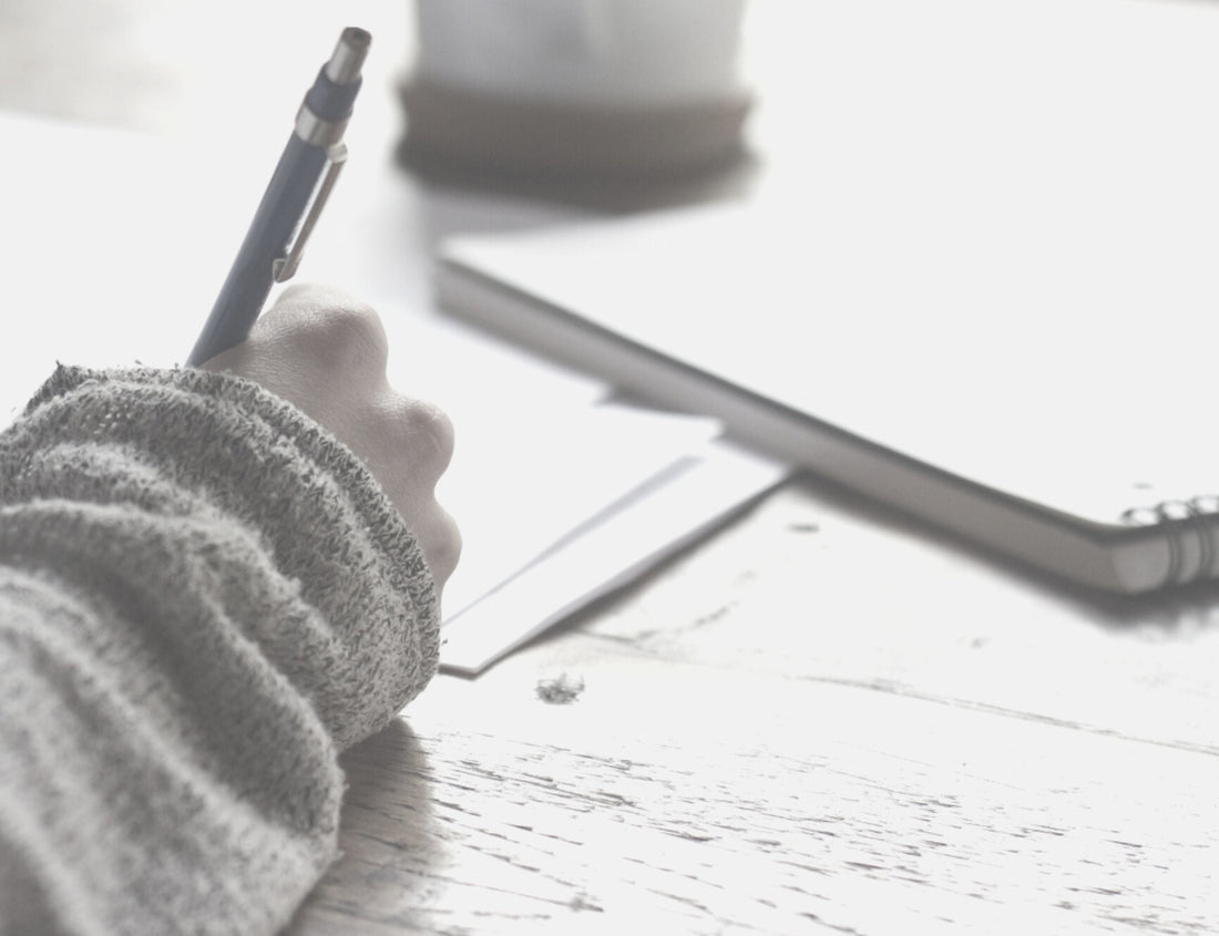 A person sitting at a table holding a pen and writing on some paper, with a coffee cup and notebaook nearby