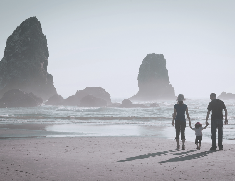 A family of 3 holding hands on a sandy shoreline with large rocks visible in the sea