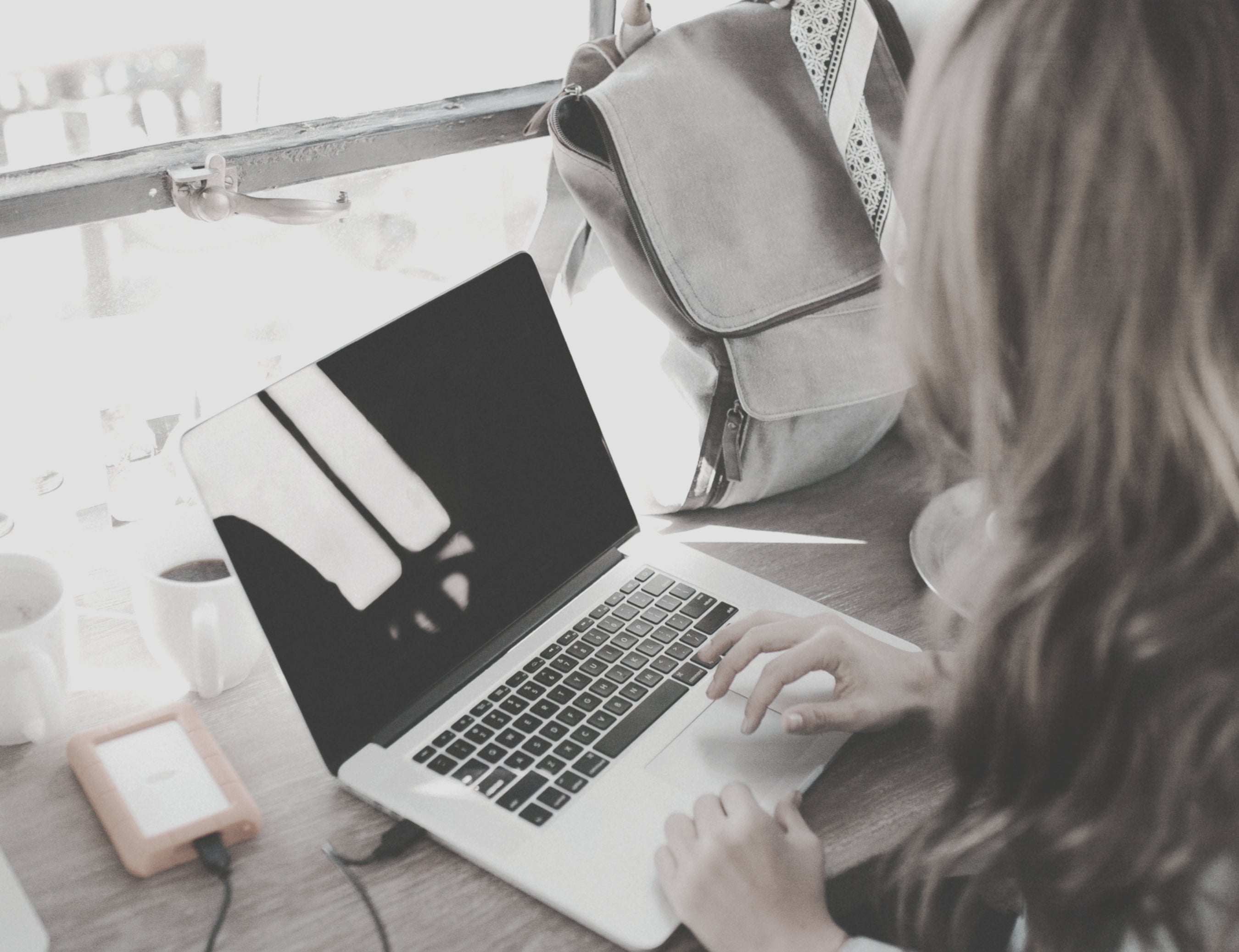 A person sitting at a laptop in a cafe window with a backback nearby