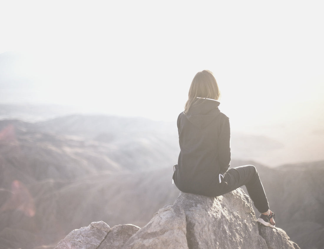 A woman sitting atop a rocky outcrop looking out across a mountain landscape