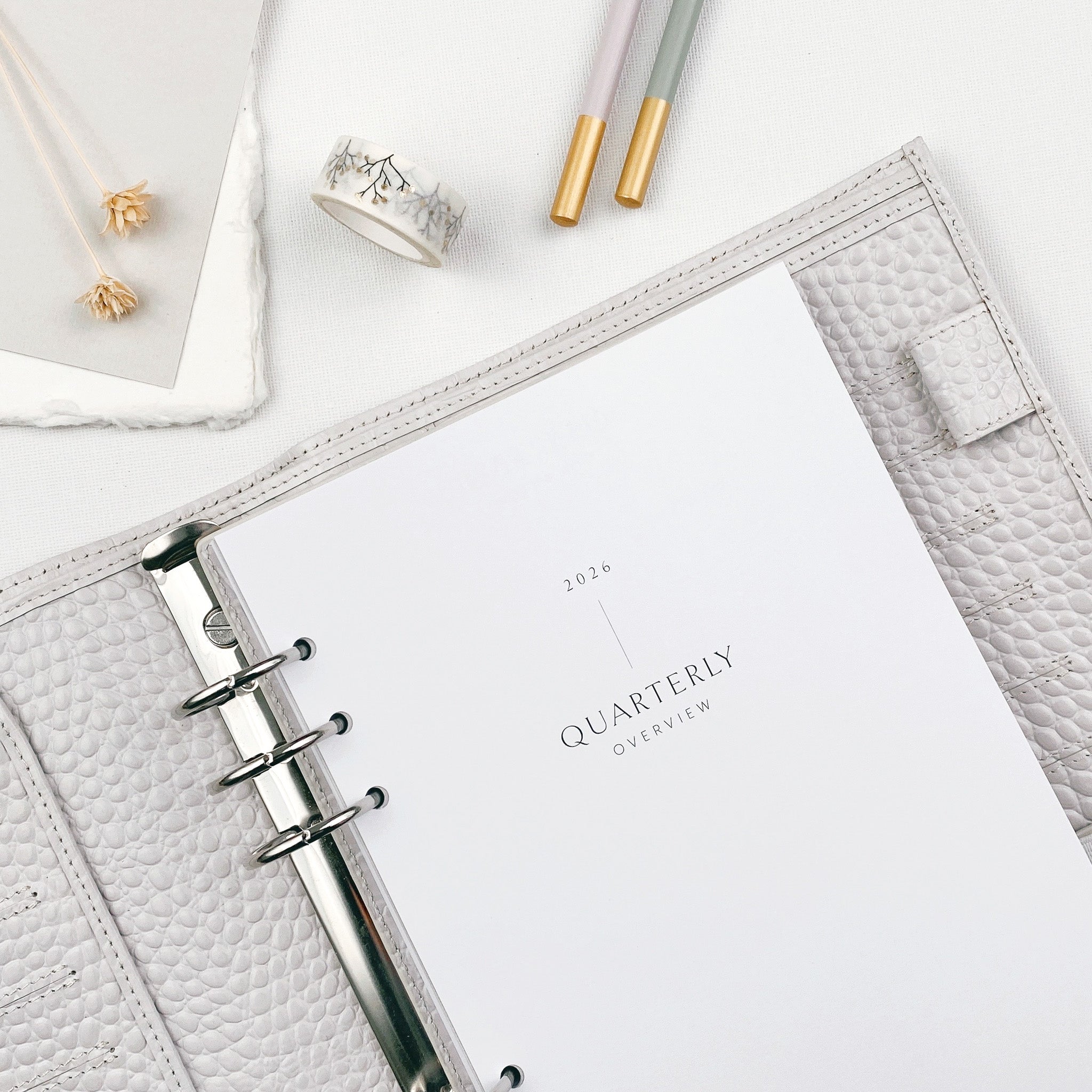 Organiser folder opened to a Quarterly Overview planner layout on a light surface with stationery items and botanicals nearby
