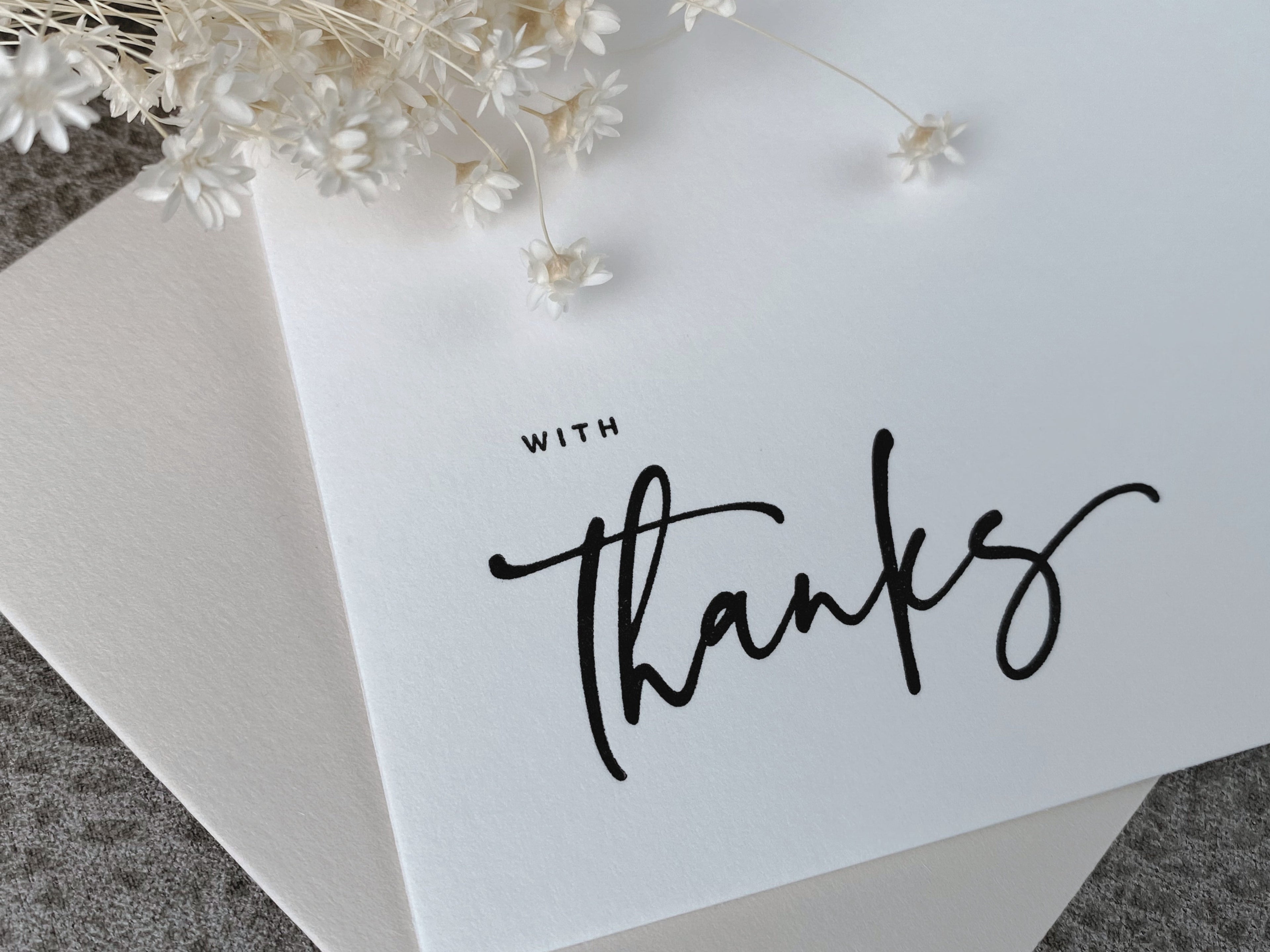 A white letterpress printed 'with thanks' greeting card and contrasting envelope set on a textured background with dried botanicals to the side