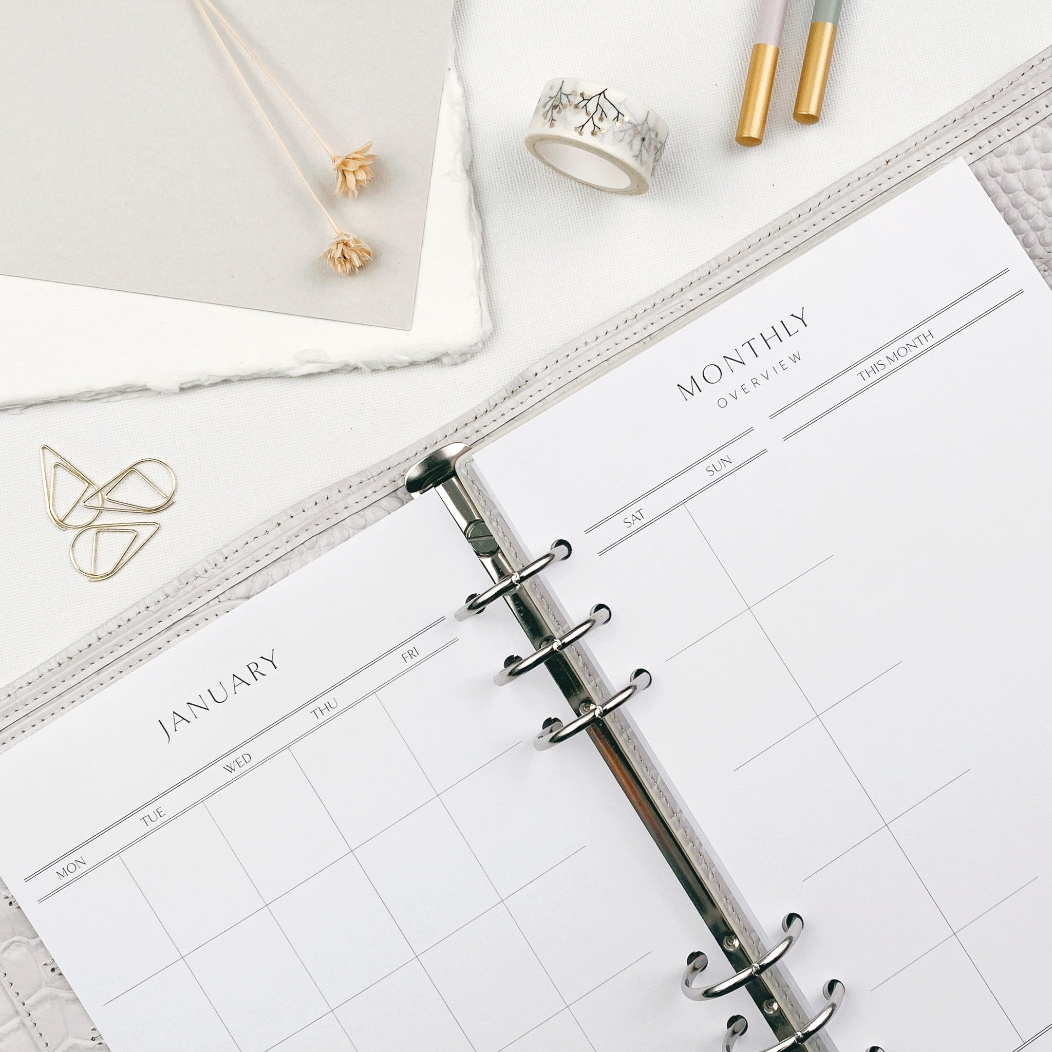 An open planner showing an undated monthly calendar grid layout, with a light desk surface and various decorative items nearby