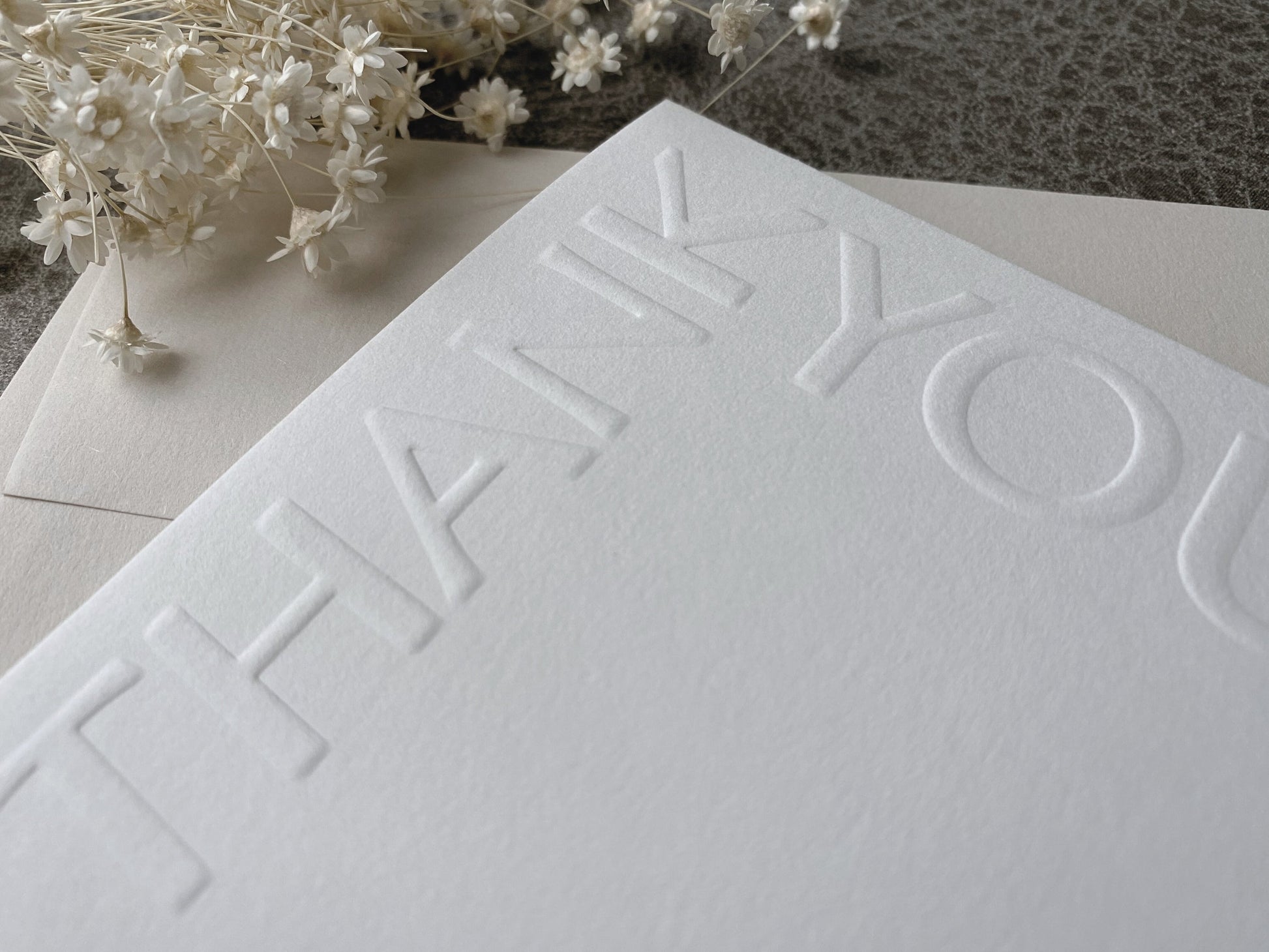 A white greeting card with 'Thank You' text embossed on the front, accompanied by a premium colored envelope, presented on a textured background with decorative flowers.