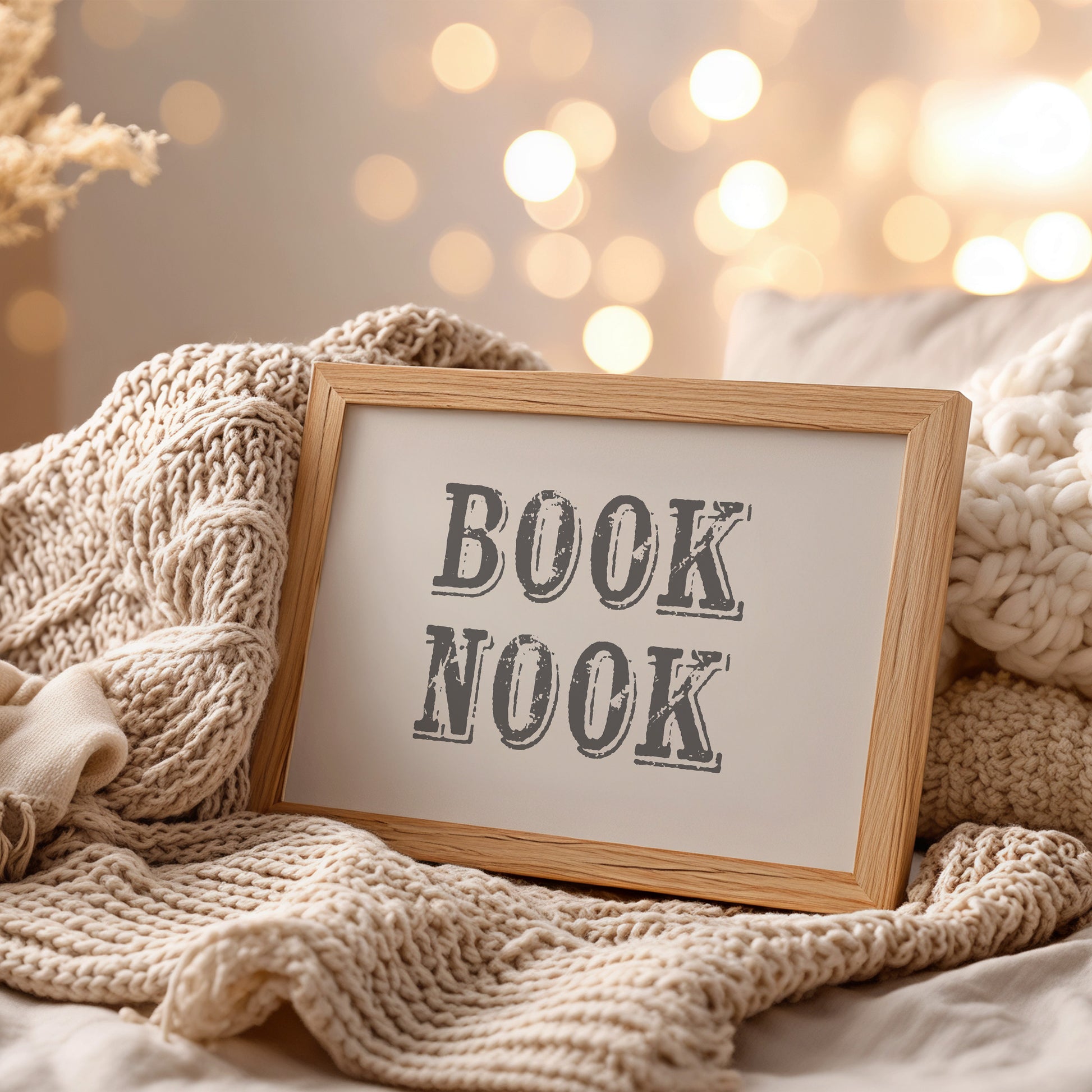 Framed art print with 'BOOK NOOK' text on a cozy blanket with blurred lights in the background