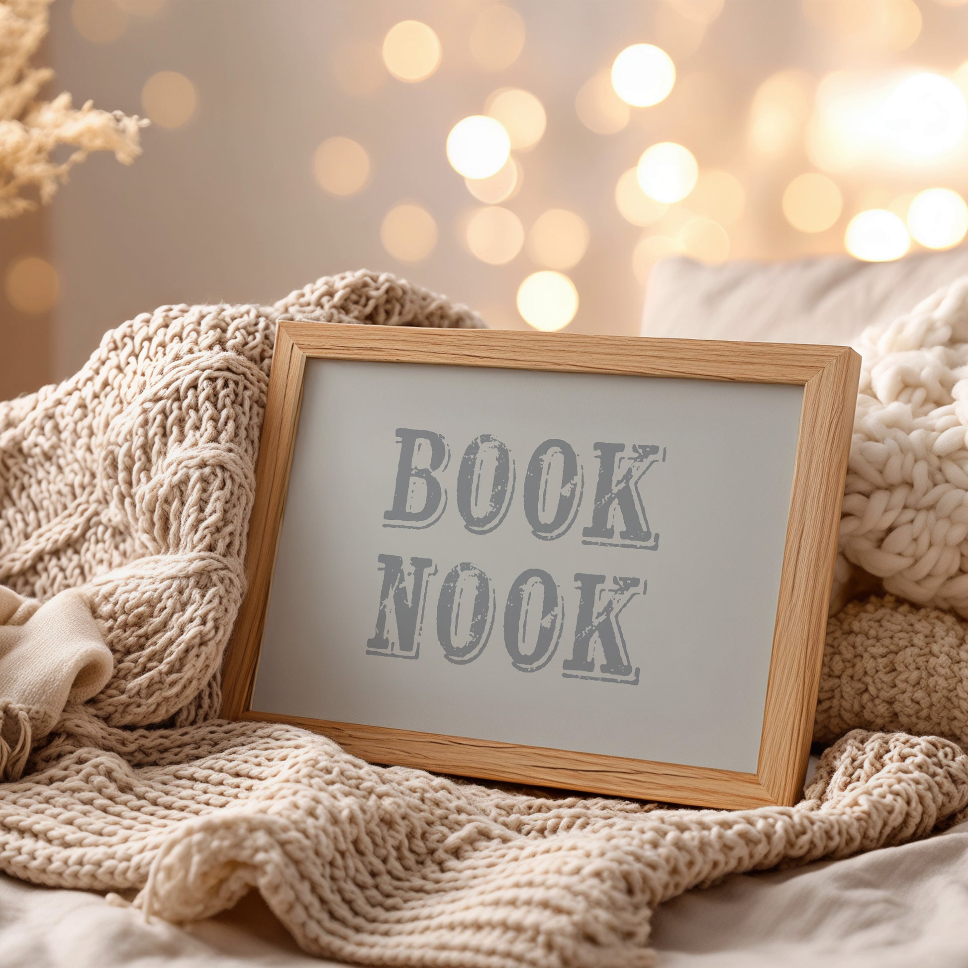 Framed wall art print with 'BOOK NOOK' text on a cozy blanket with blurred lights in the background