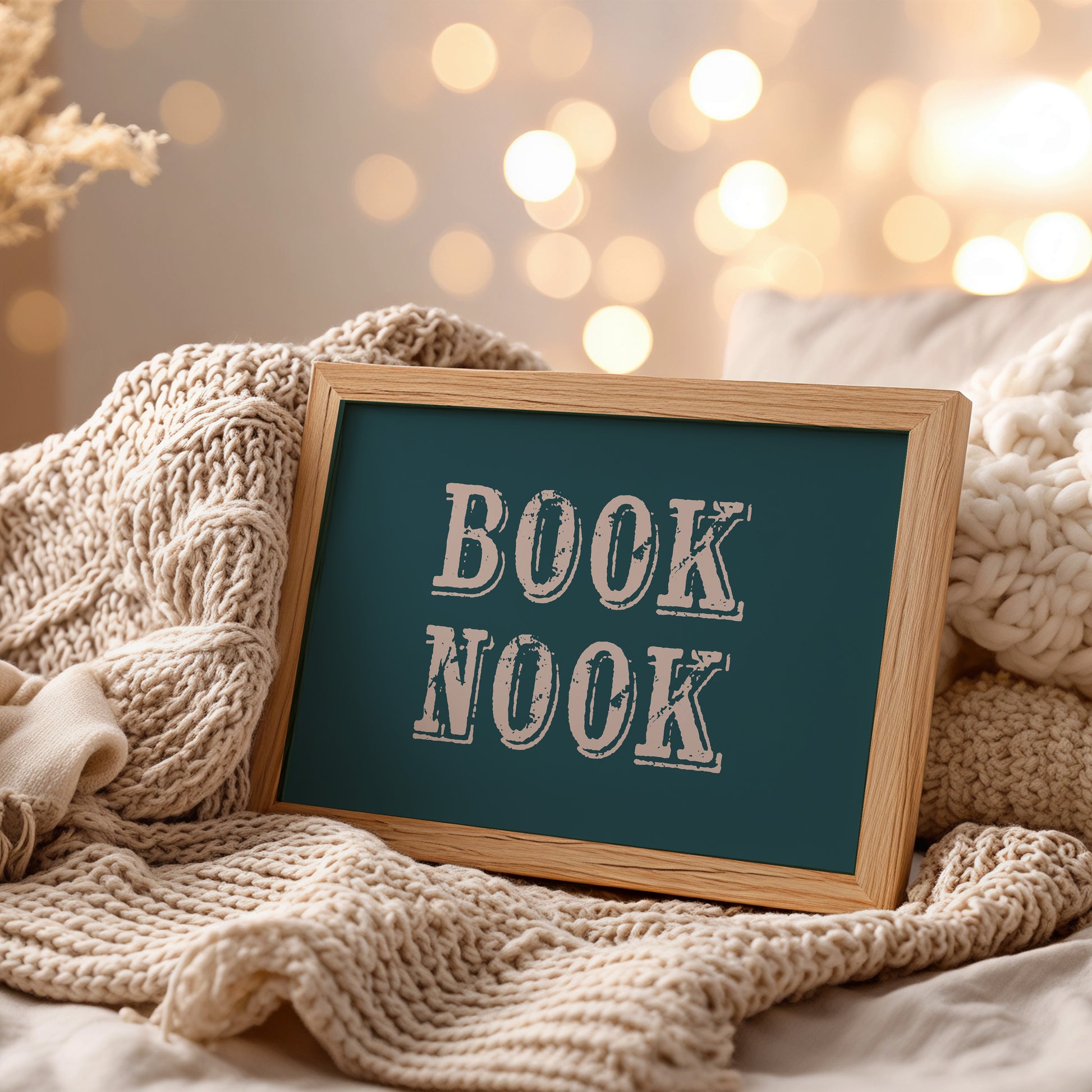 Framed wall art with 'BOOK NOOK' text on a cozy blanket with blurred lights in the background