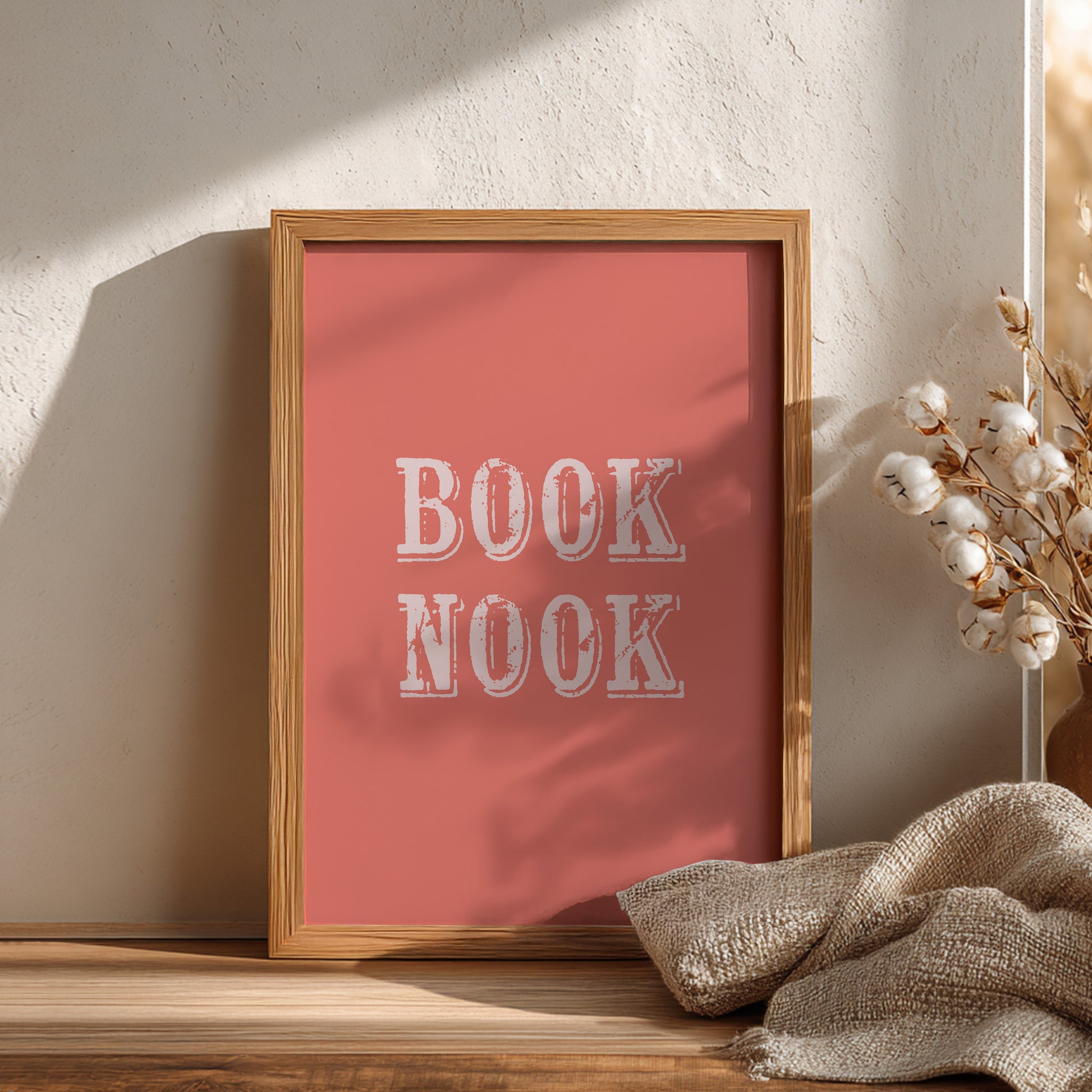 Framed poster with 'BOOK NOOK' text on a coral pink background, placed on a wooden surface with decorative elements.
