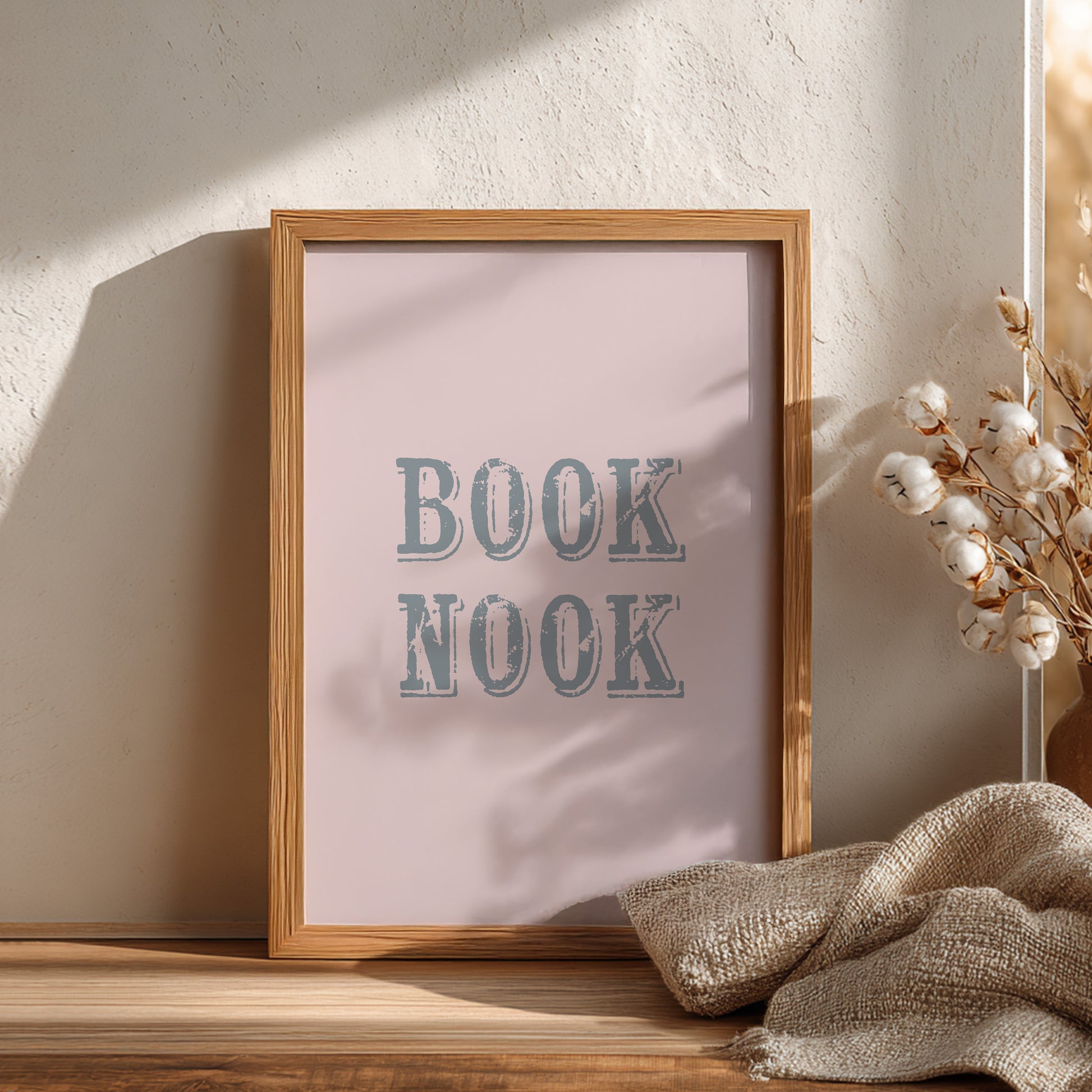 Framed artwork with 'BOOK NOOK' text on a light pink background, placed on a wooden surface with decorative elements.