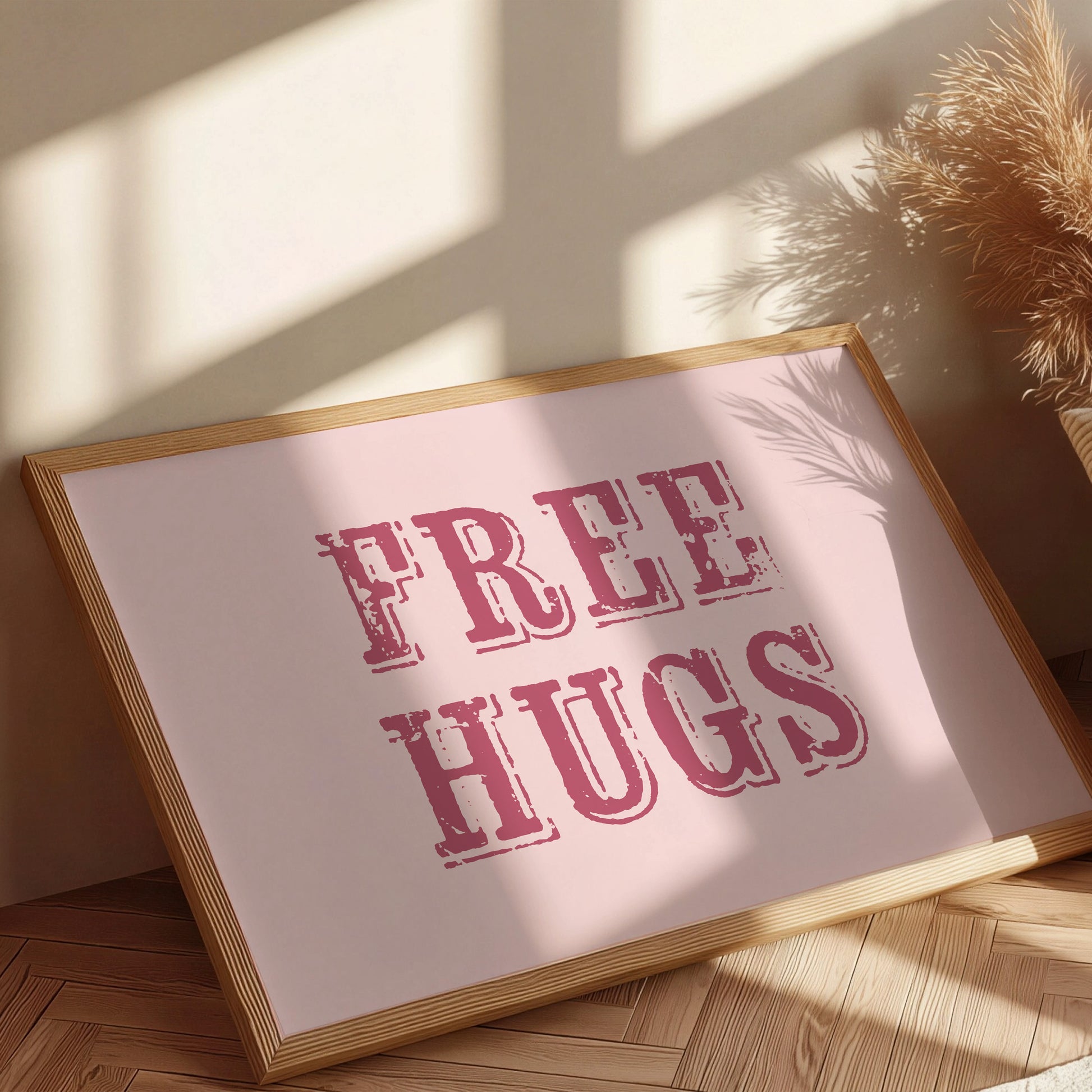 Framed artwork with 'FREE HUGS' text on a pink background, placed on a wooden floor with sunlight casting shadows.