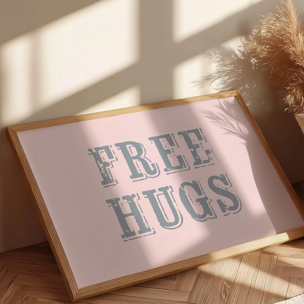 Framed sign with 'FREE HUGS' text on a pink background, placed on a wooden floor with sunlight casting shadows.