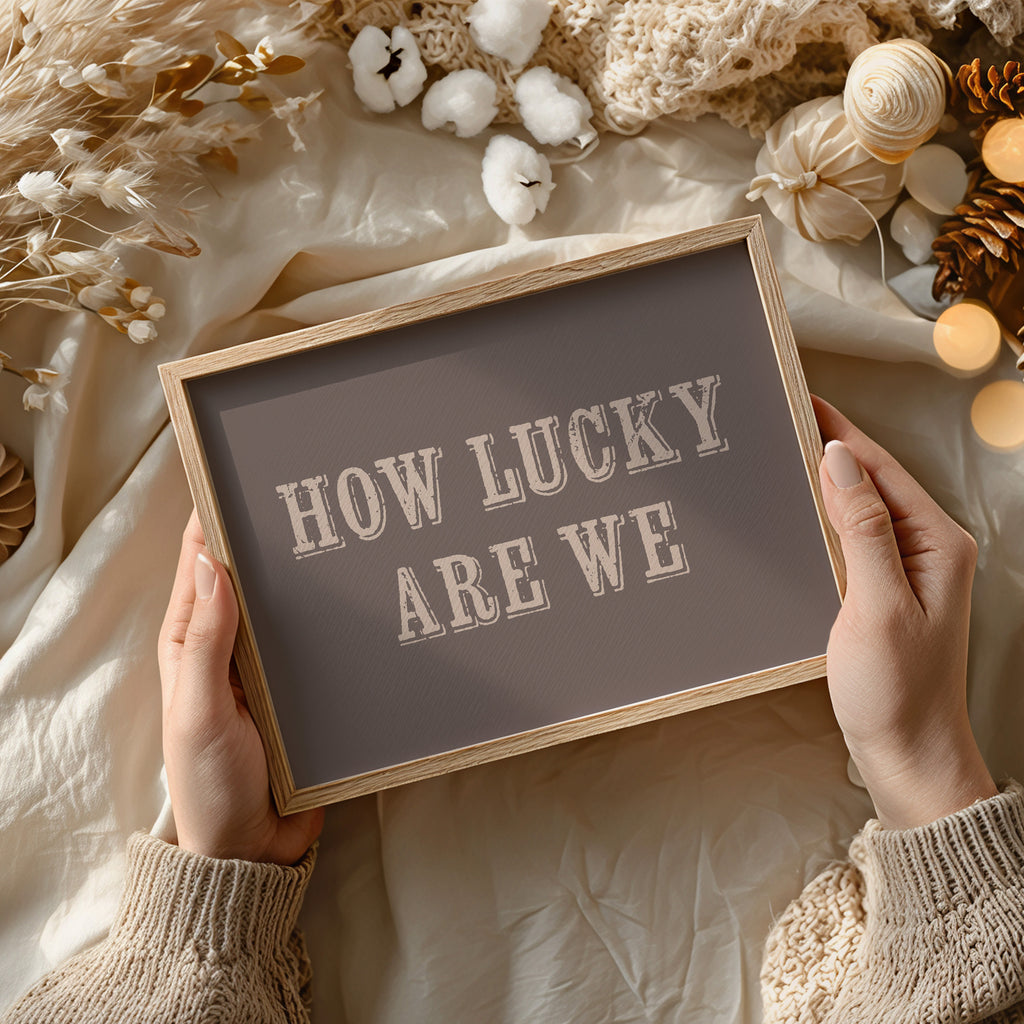 Framed print with 'HOW LUCKY ARE WE' text held by hands against a soft, decorative background.