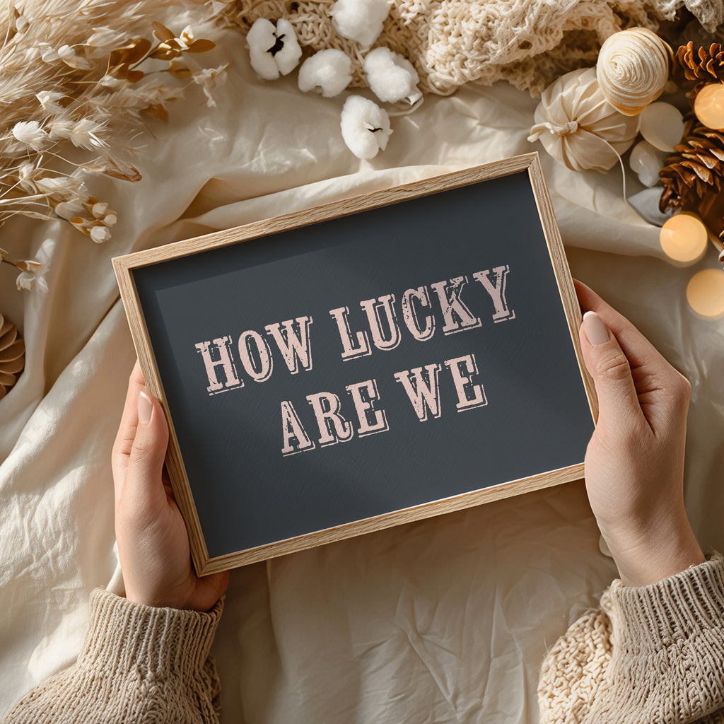 Person holding a framed art print with 'HOW LUCKY ARE WE' text against a decorative background.