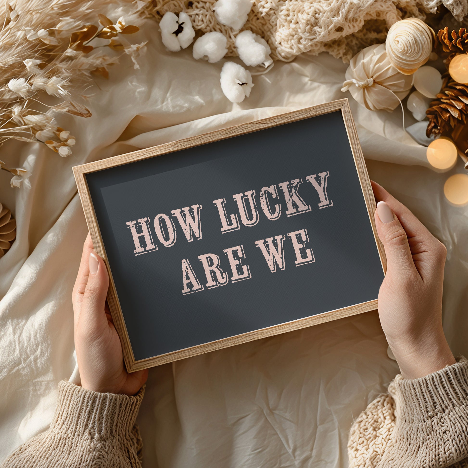 Person holding a framed art print with 'HOW LUCKY ARE WE' text against a decorative background.