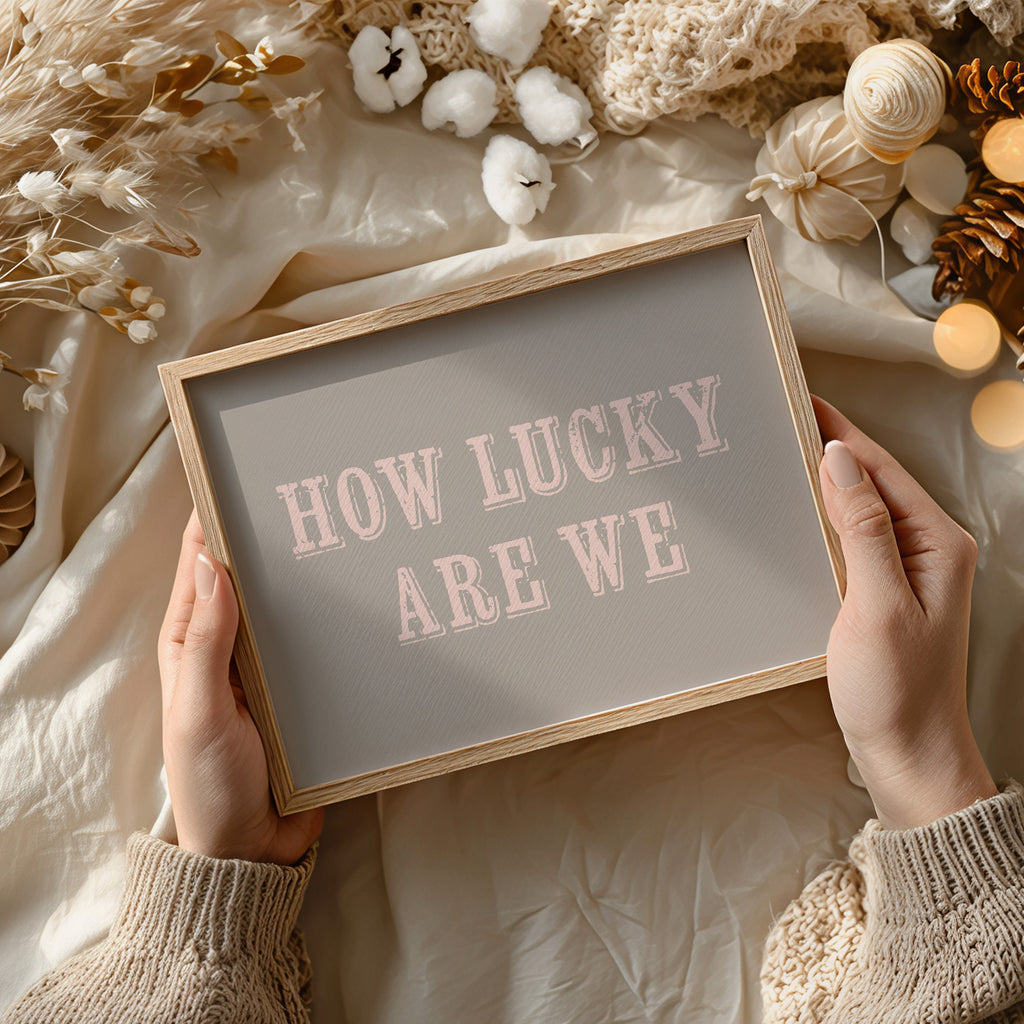 Framed print with 'HOW LUCKY ARE WE' text held by hands against a soft, decorative background.
