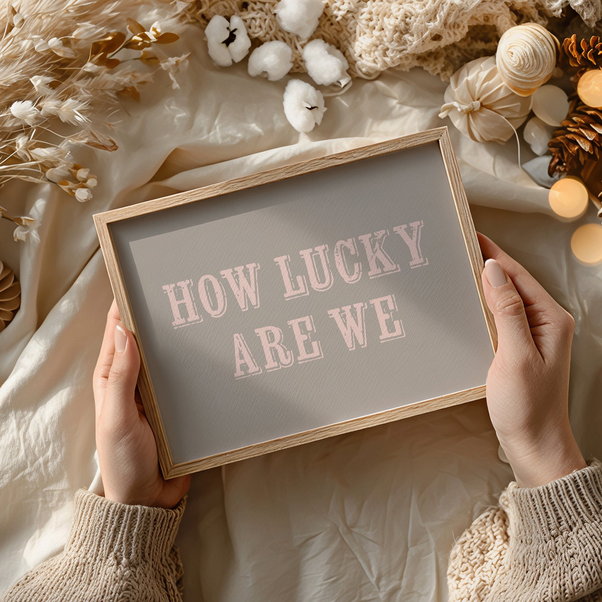 Framed print with 'HOW LUCKY ARE WE' text held by hands against a soft, decorative background.