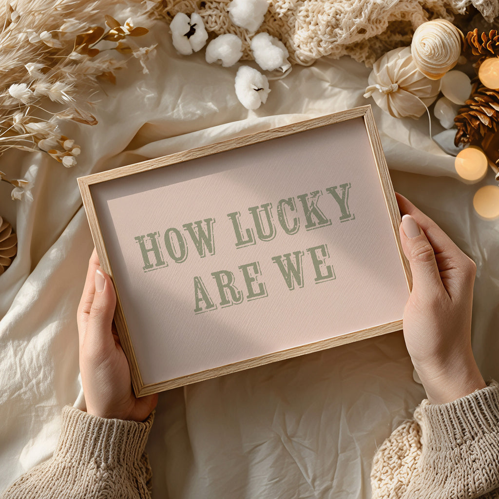 Framed sign with 'HOW LUCKY ARE WE' text held by hands against a soft, decorative background.