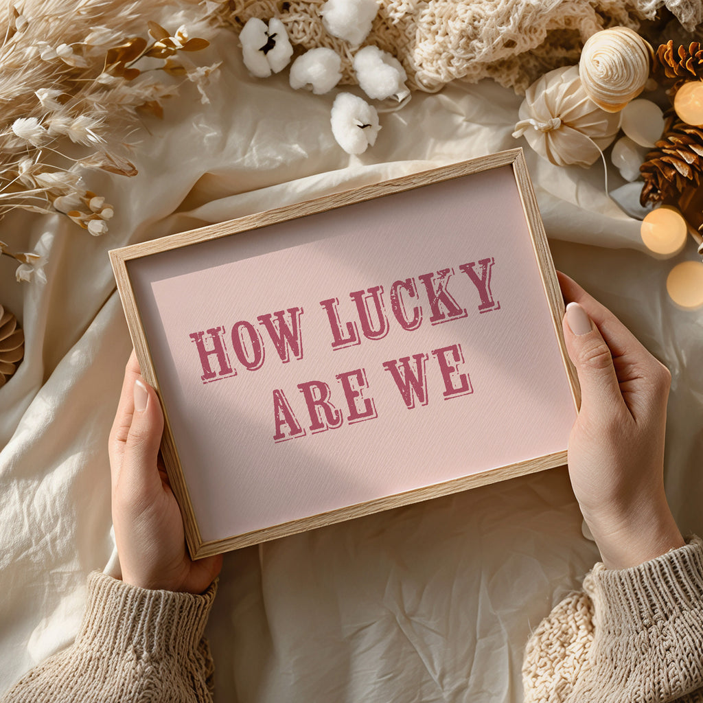 Framed art print with 'HOW LUCKY ARE WE' text held by hands against a decorative background.