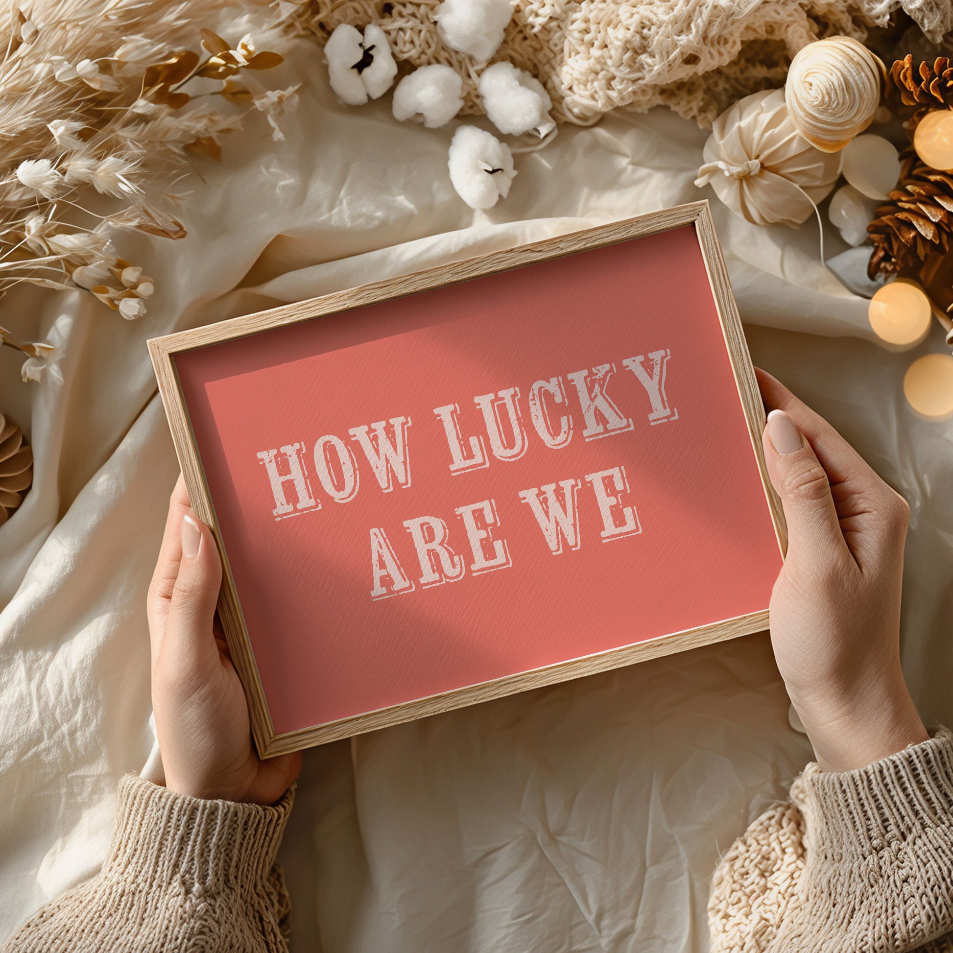 Framed art print with 'HOW LUCKY ARE WE' text held by hands against a decorative background.