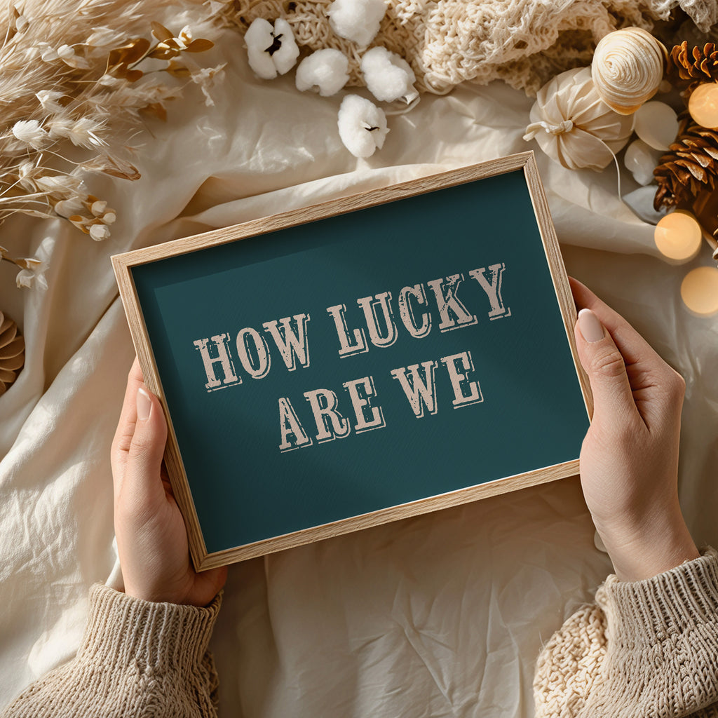 Person holding a framed print with 'HOW LUCKY ARE WE' text against a decorative background.