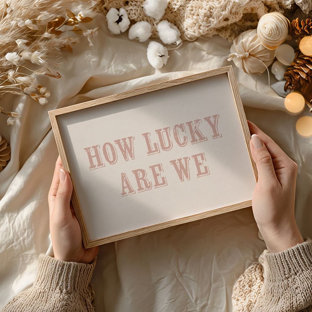 Framed sign with 'HOW LUCKY ARE WE' text held by hands against a soft, decorative background.
