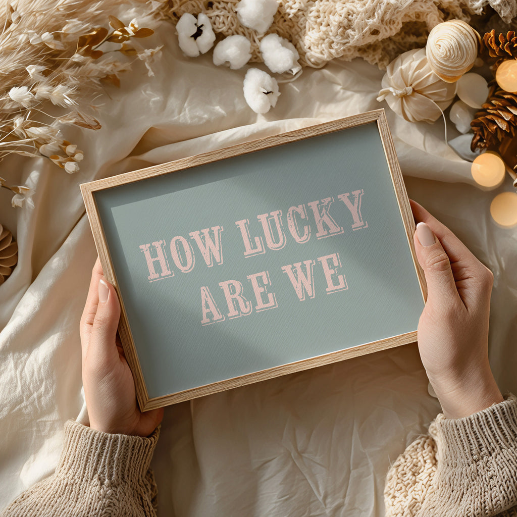 Framed art print with 'HOW LUCKY ARE WE' text held by hands against a decorative background.
