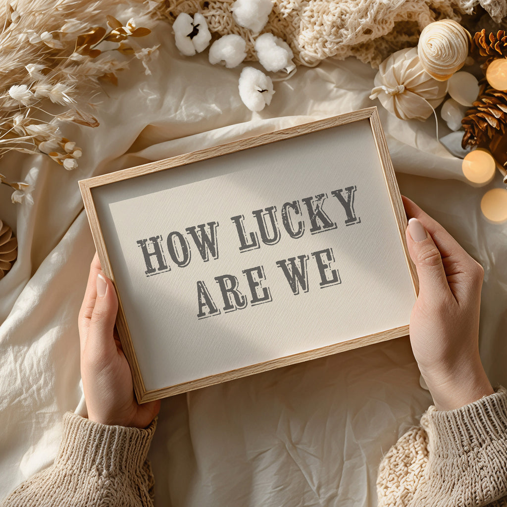 Framed print with 'HOW LUCKY ARE WE' text held by hands against a soft, decorative background.