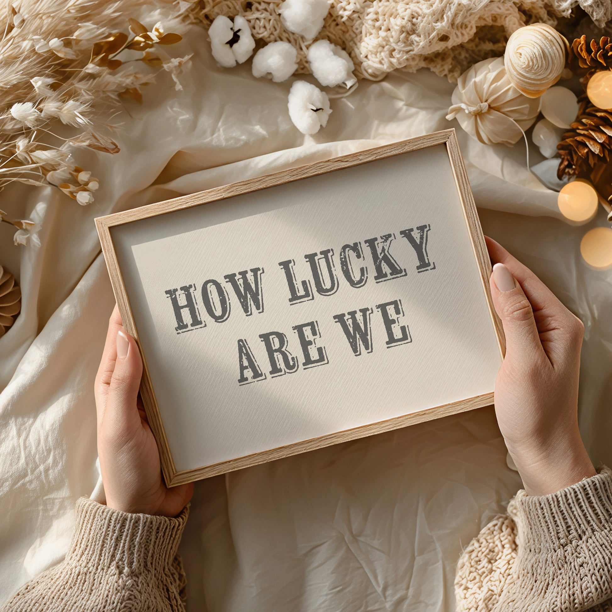 Framed print with 'HOW LUCKY ARE WE' text held by hands against a soft, decorative background.