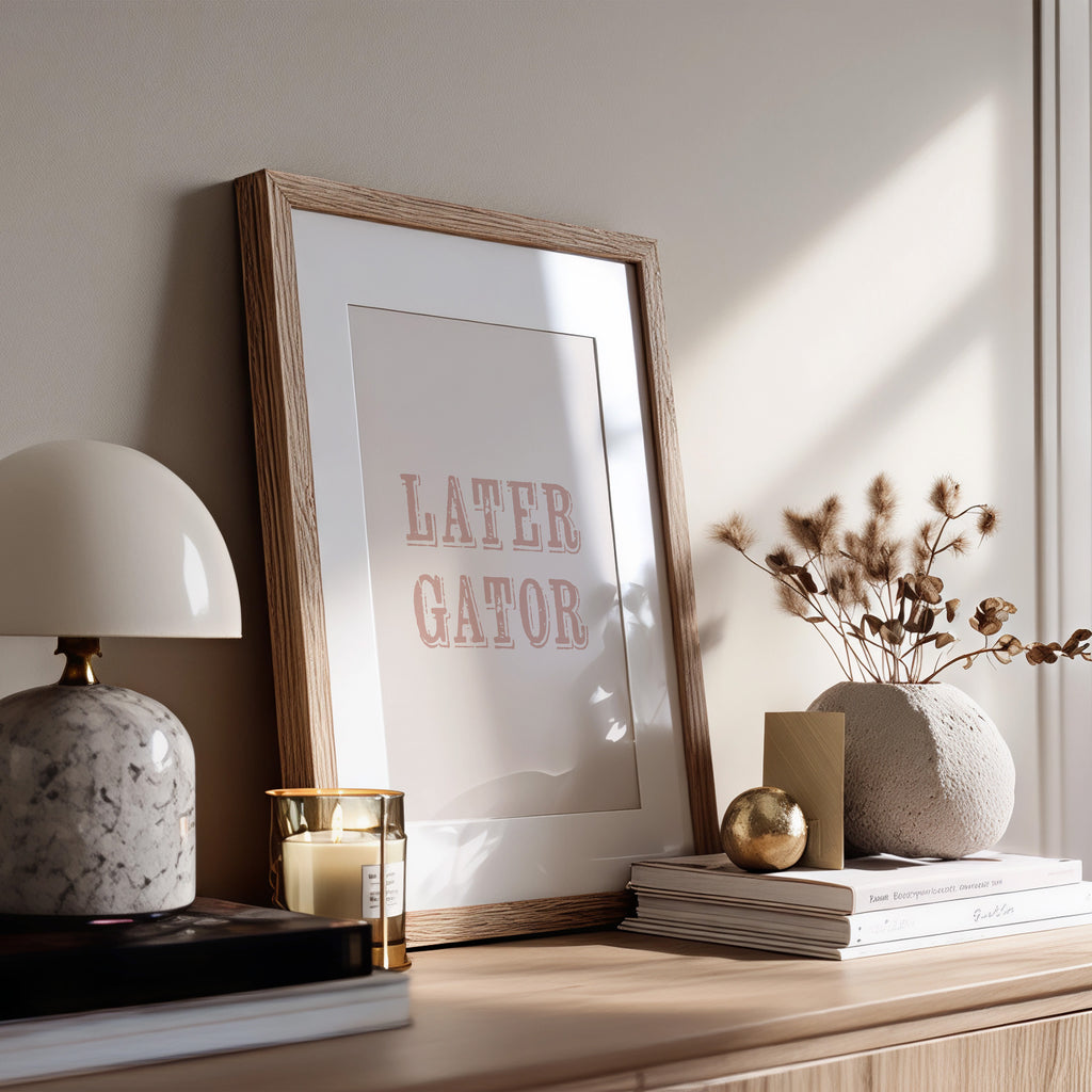 Framed print with 'LATER GATOR' text on a wooden surface with decor items.