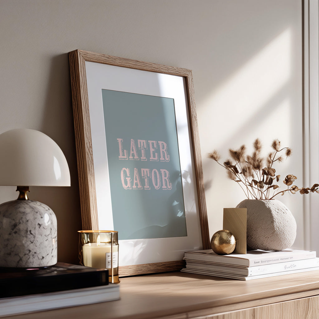 Framed print with 'LATER, GATOR' text on a wooden surface with decor items.