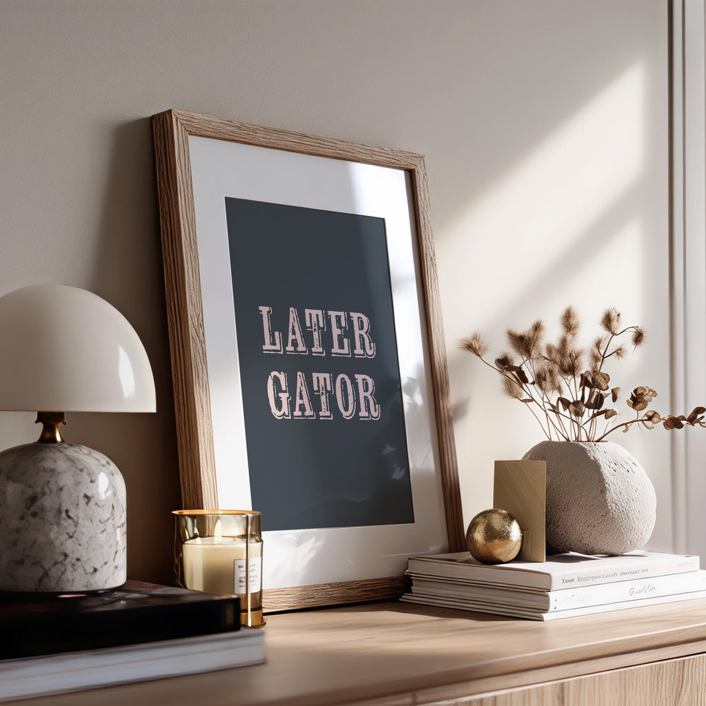 Framed print with 'LATER GATOR' text on a wooden surface with decor items.