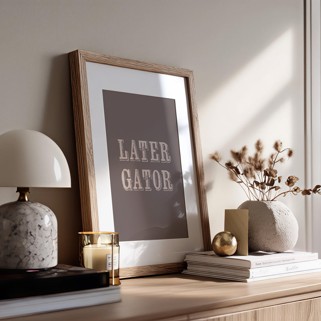Framed print with 'LATER GATOR' text on a wooden surface with decor items.