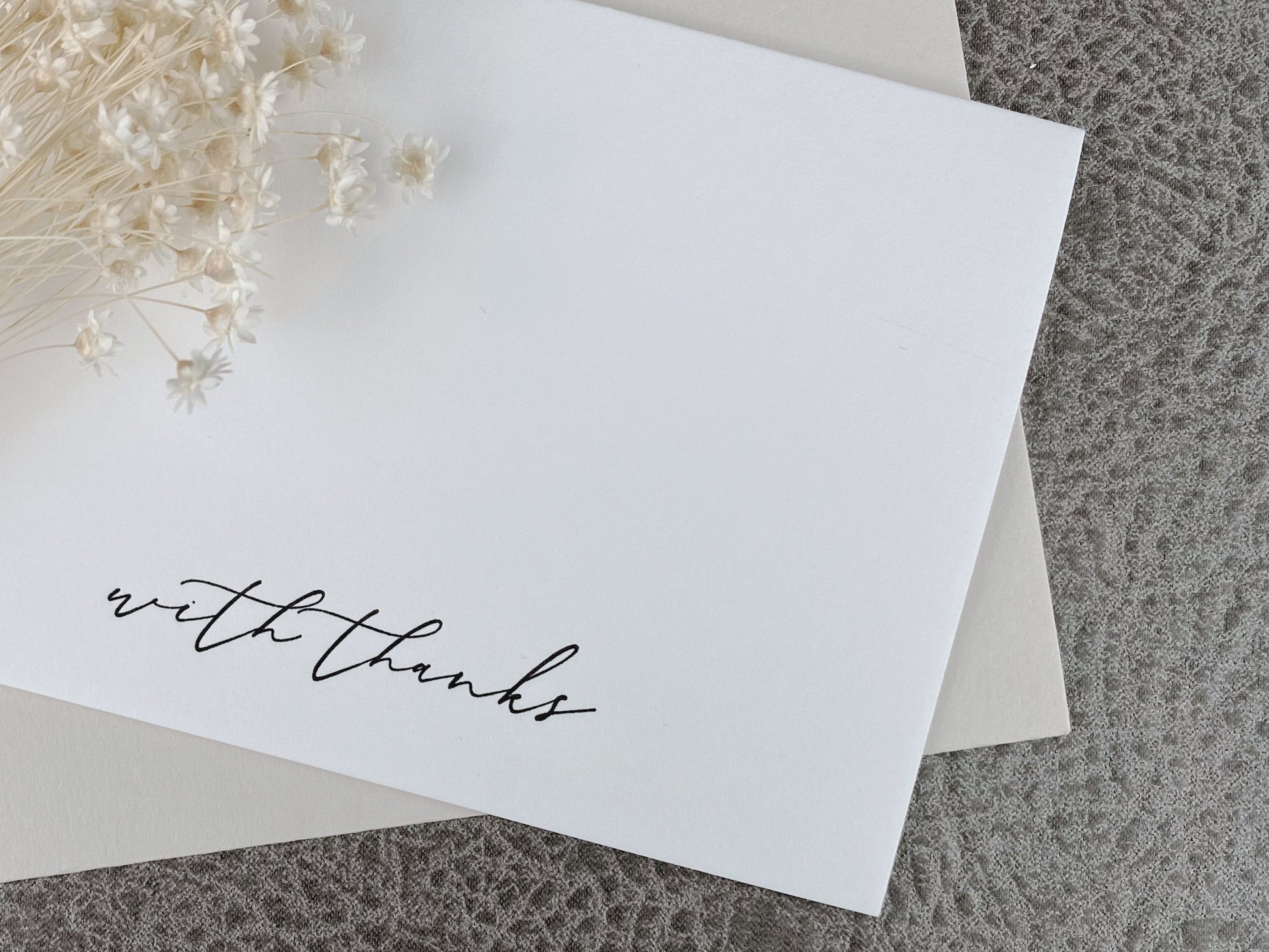 A white letterpress 'With Thanks' greeting card displayed with a premium envelope, resting on a textured background with decorative dried flowers on the side.