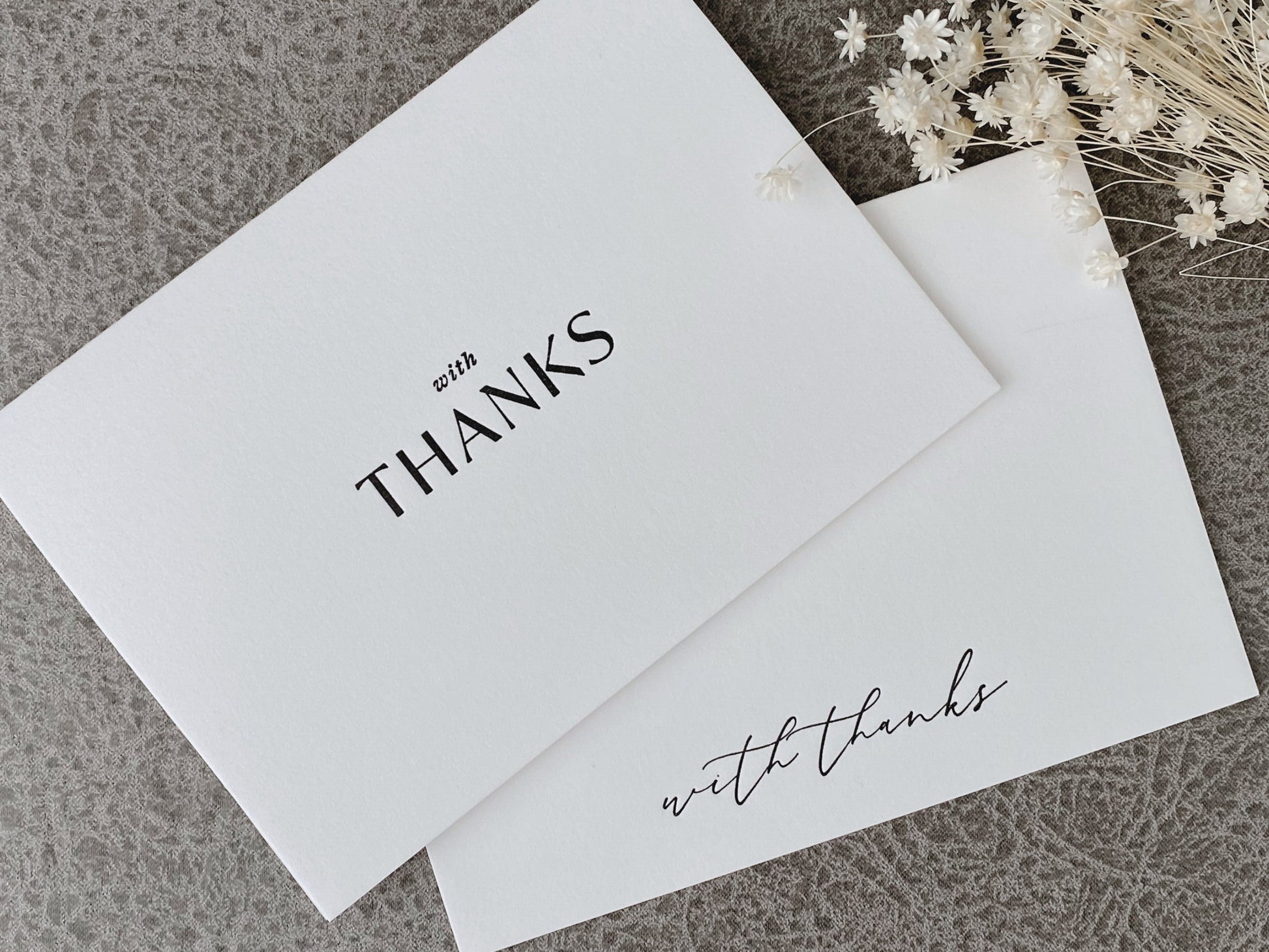 A set of white letterpress thank you greeting cards with the text 'With Thanks' printed in black. The cards are displayed with a floral decoration in the background.