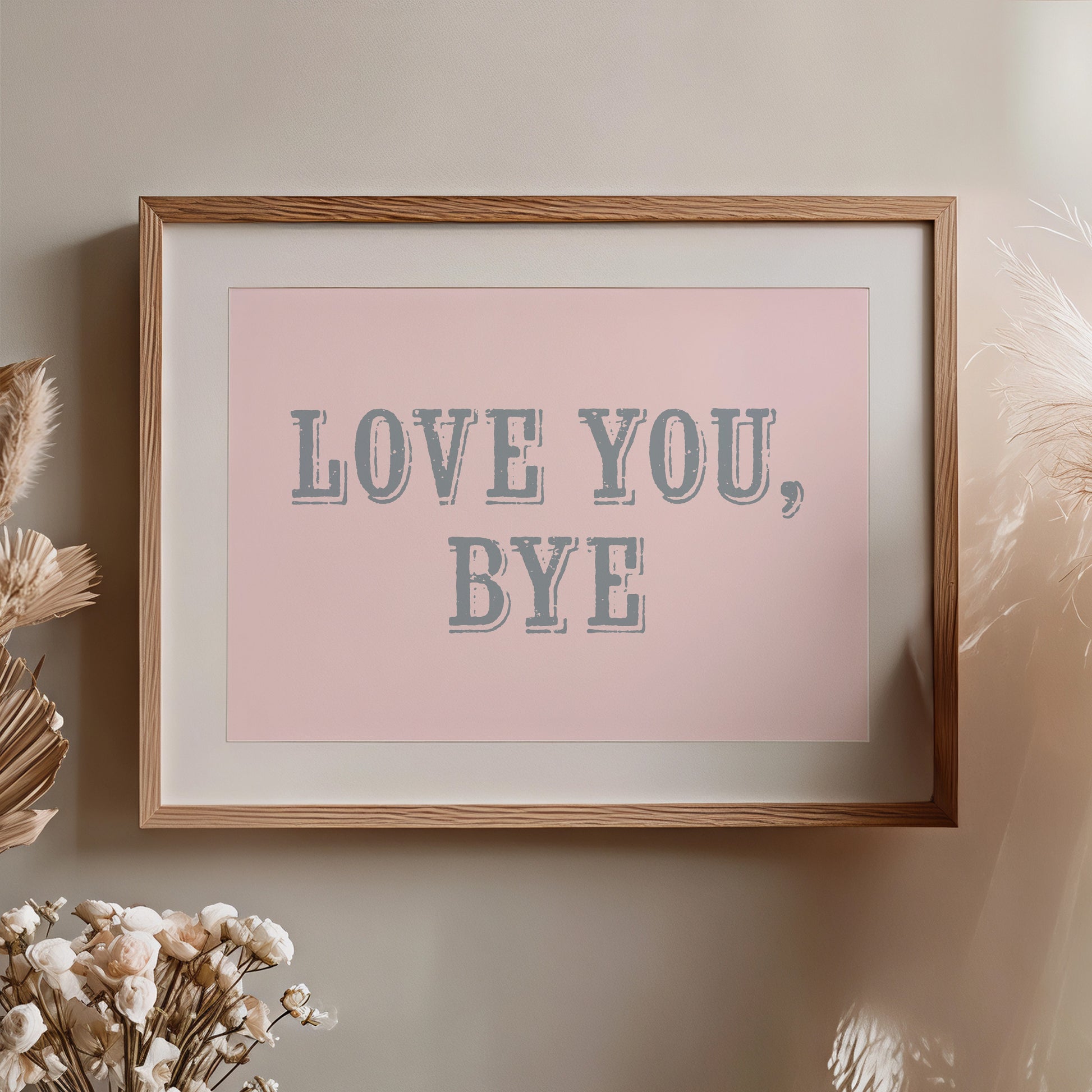 Framed artwork with 'LOVE YOU, BYE' text on a pink background, placed on a wall with decorative elements nearby.