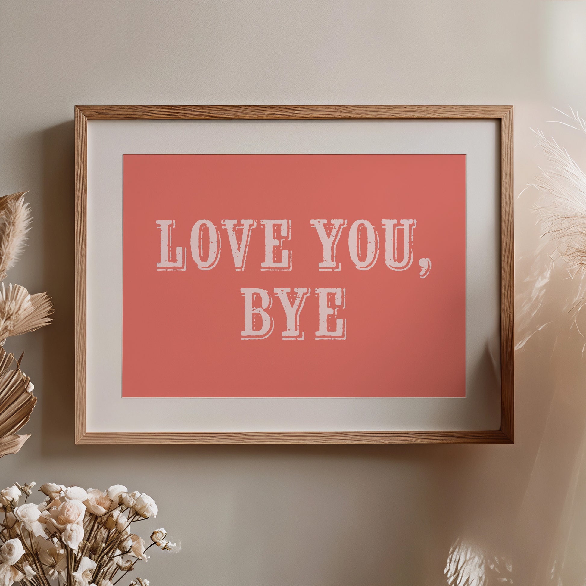 Framed artwork with 'LOVE YOU, BYE' text on a coral background, hung on a wall with decorative elements nearby.