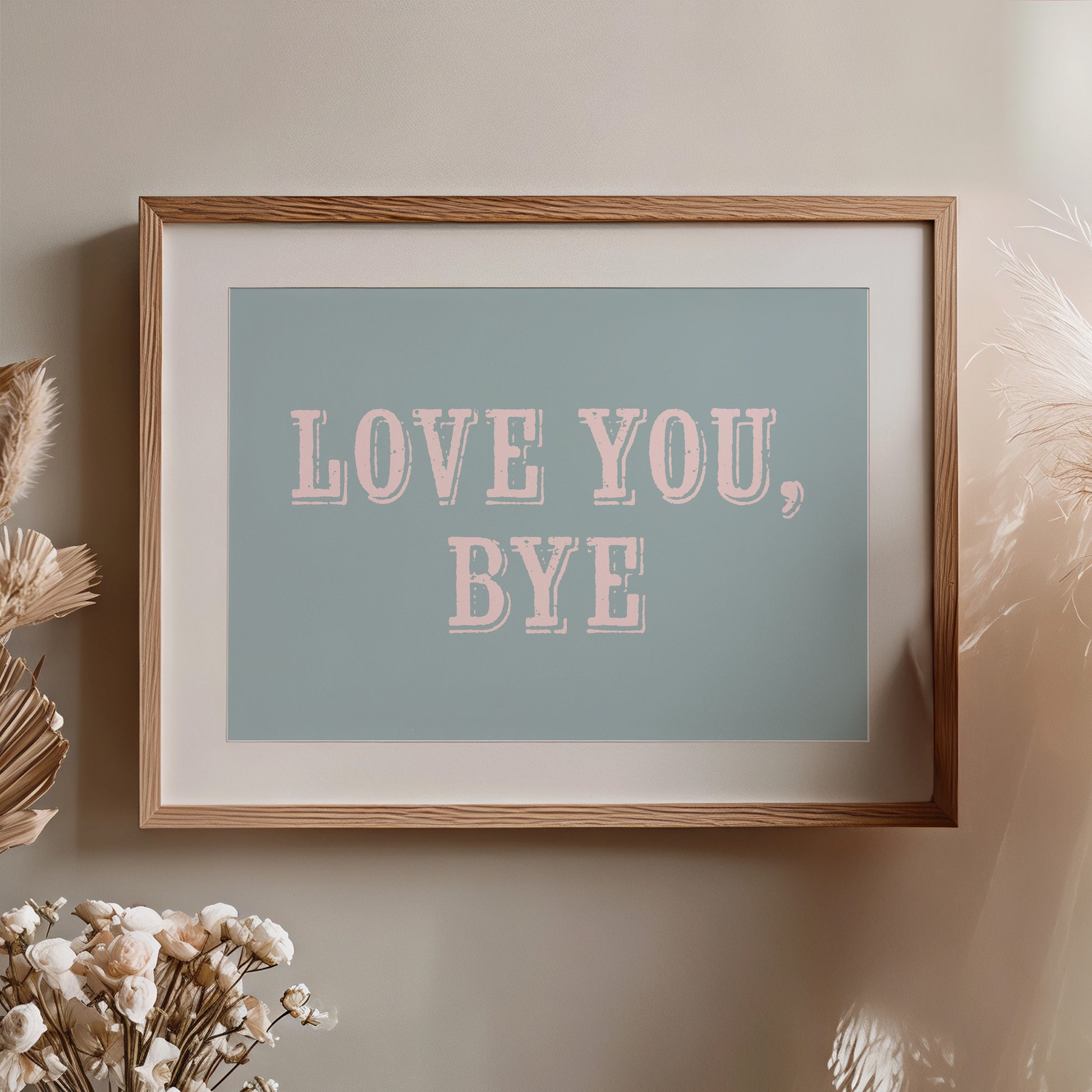 Framed artwork with 'LOVE YOU, BYE' text on a light blue background, placed on a wall with decorative elements nearby.