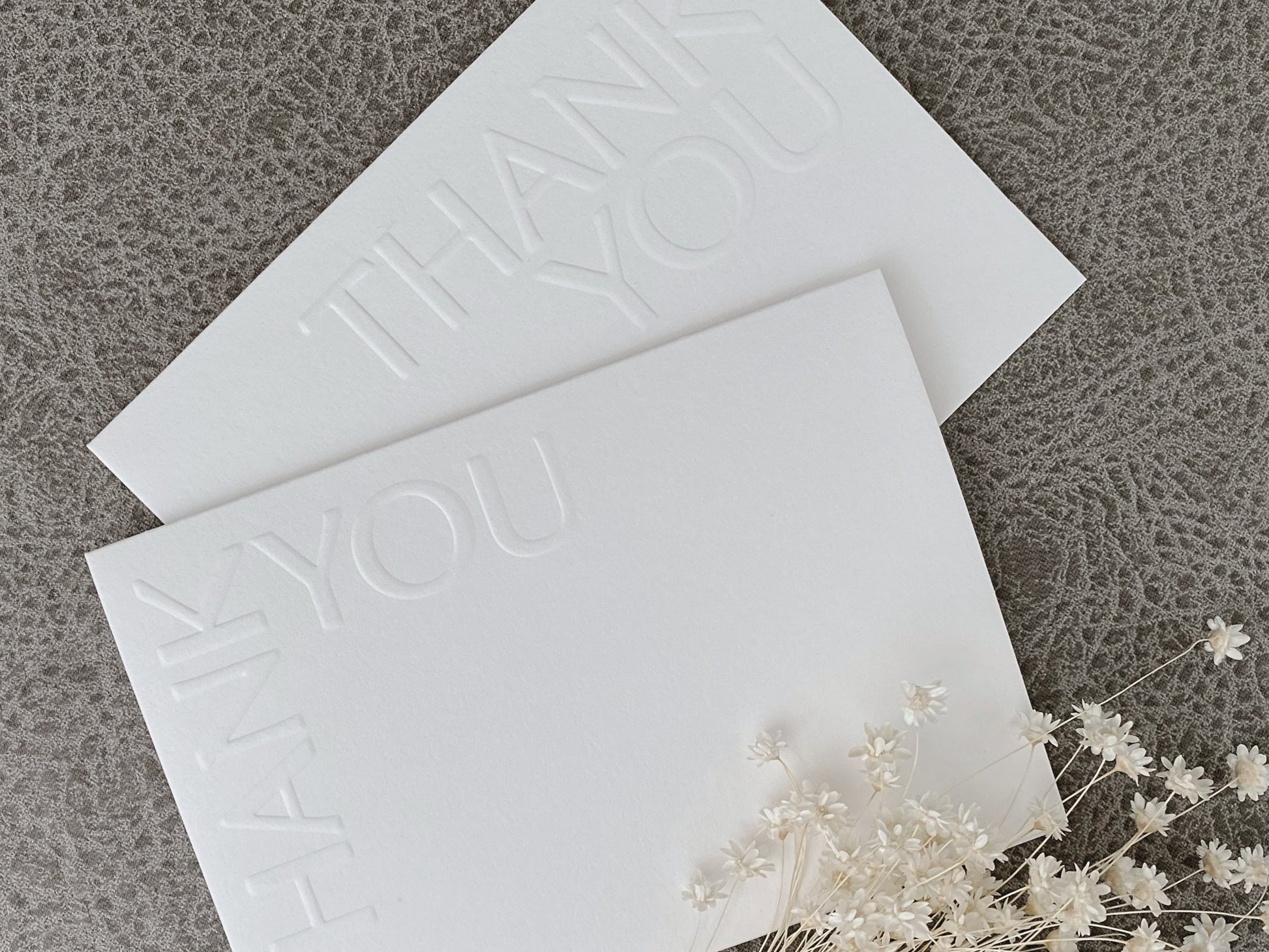 A set of two white embossed thank you greeting cards with 'Thank You' text, presented on a textured background with a floral arrangement to the side.