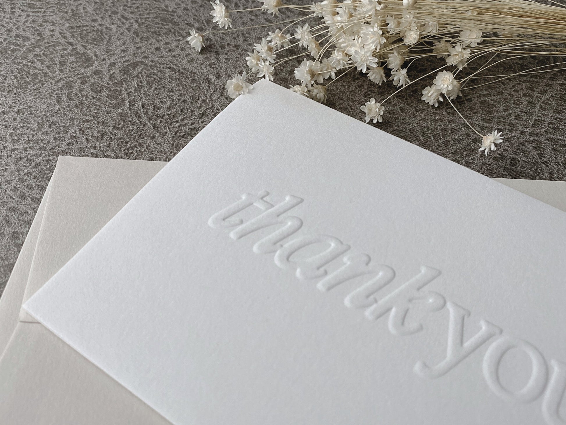 A white greeting card with the words 'Thank You' embossed on the front, accompanied by a premium colored envelope.
