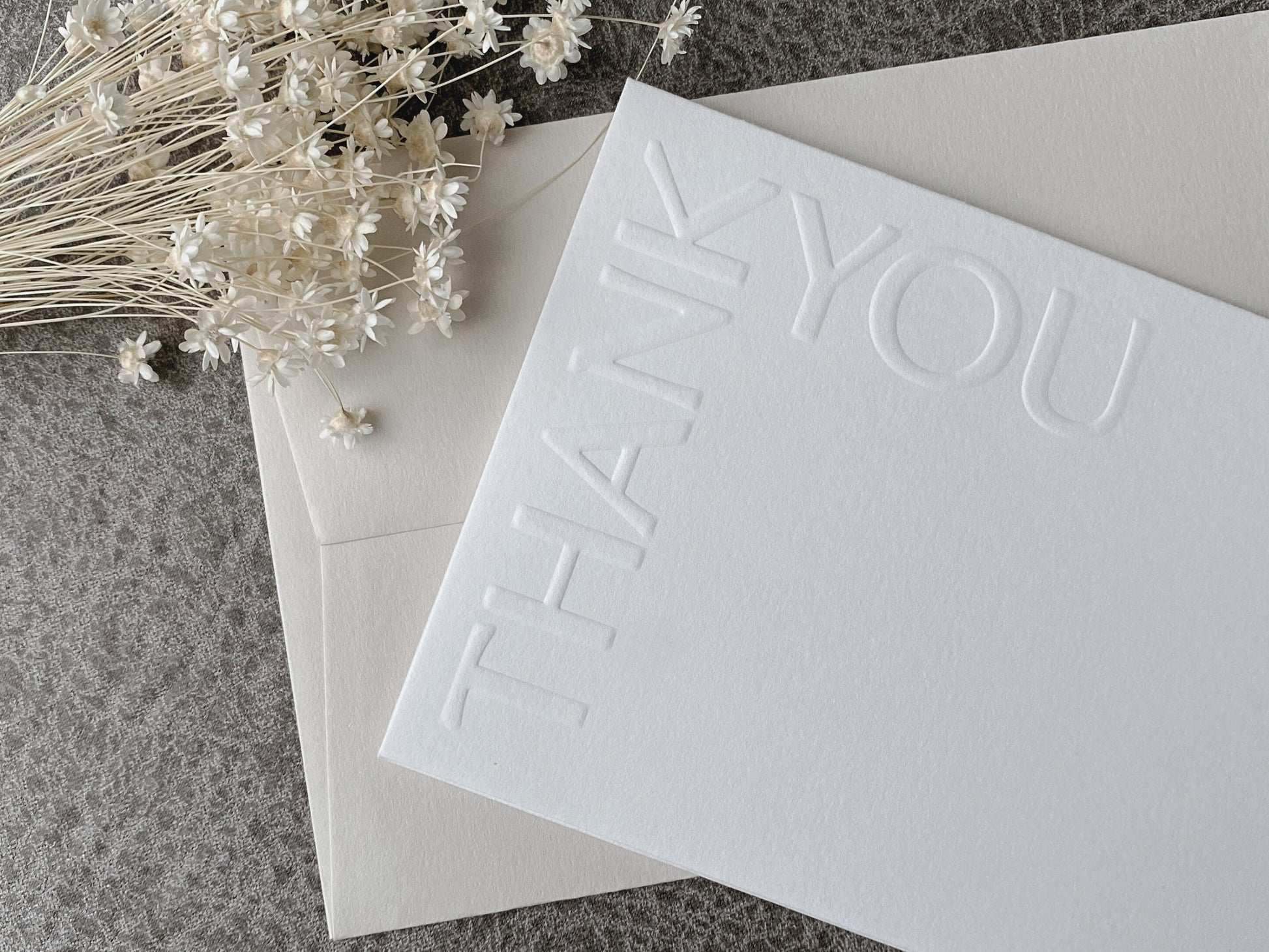 A white greeting card with 'Thank You' text embossed on the front, accompanied by a premium colored envelope, presented on a textured background with decorative flowers.