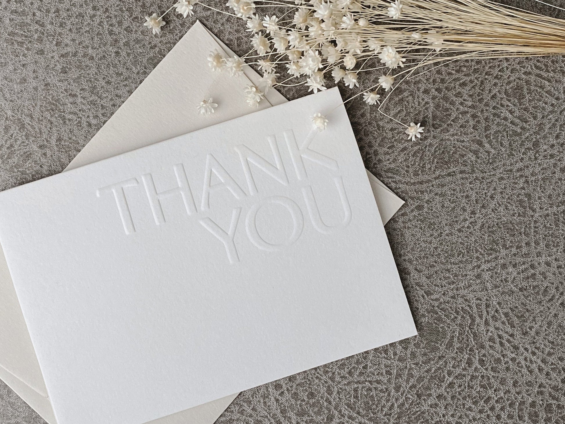 A minimalist embossed 'THANK YOU' greeting card in white, placed on a textured surface with a dried botanical decoration to the side.