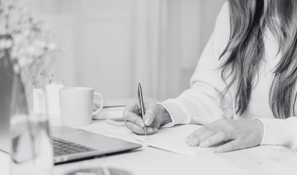 Person sitting at a desk with a laptop and a cup, writing with a pen.