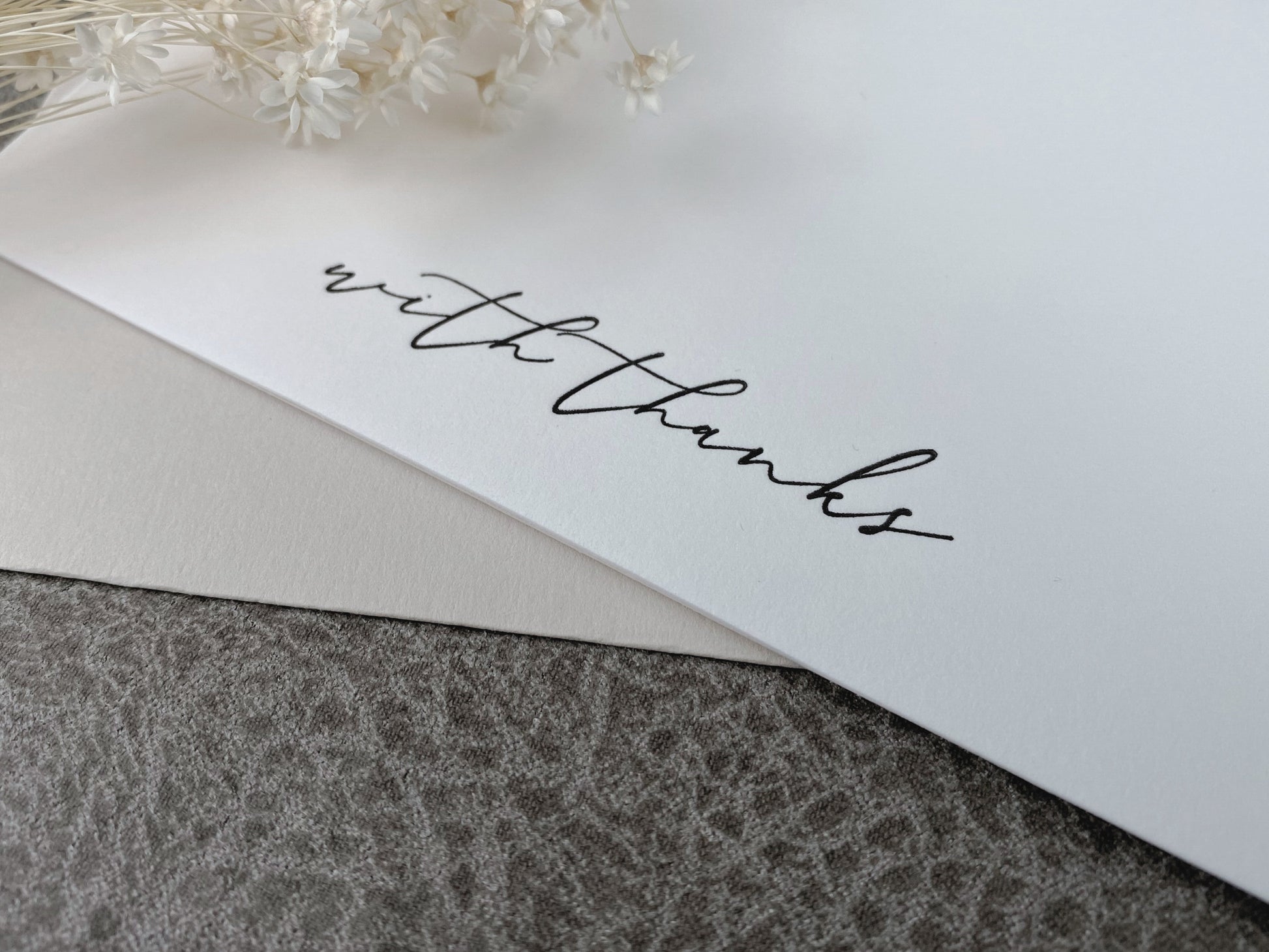A white letterpress 'With Thanks' greeting card displayed with a premium envelope, resting on a textured background with decorative dried flowers on the side.