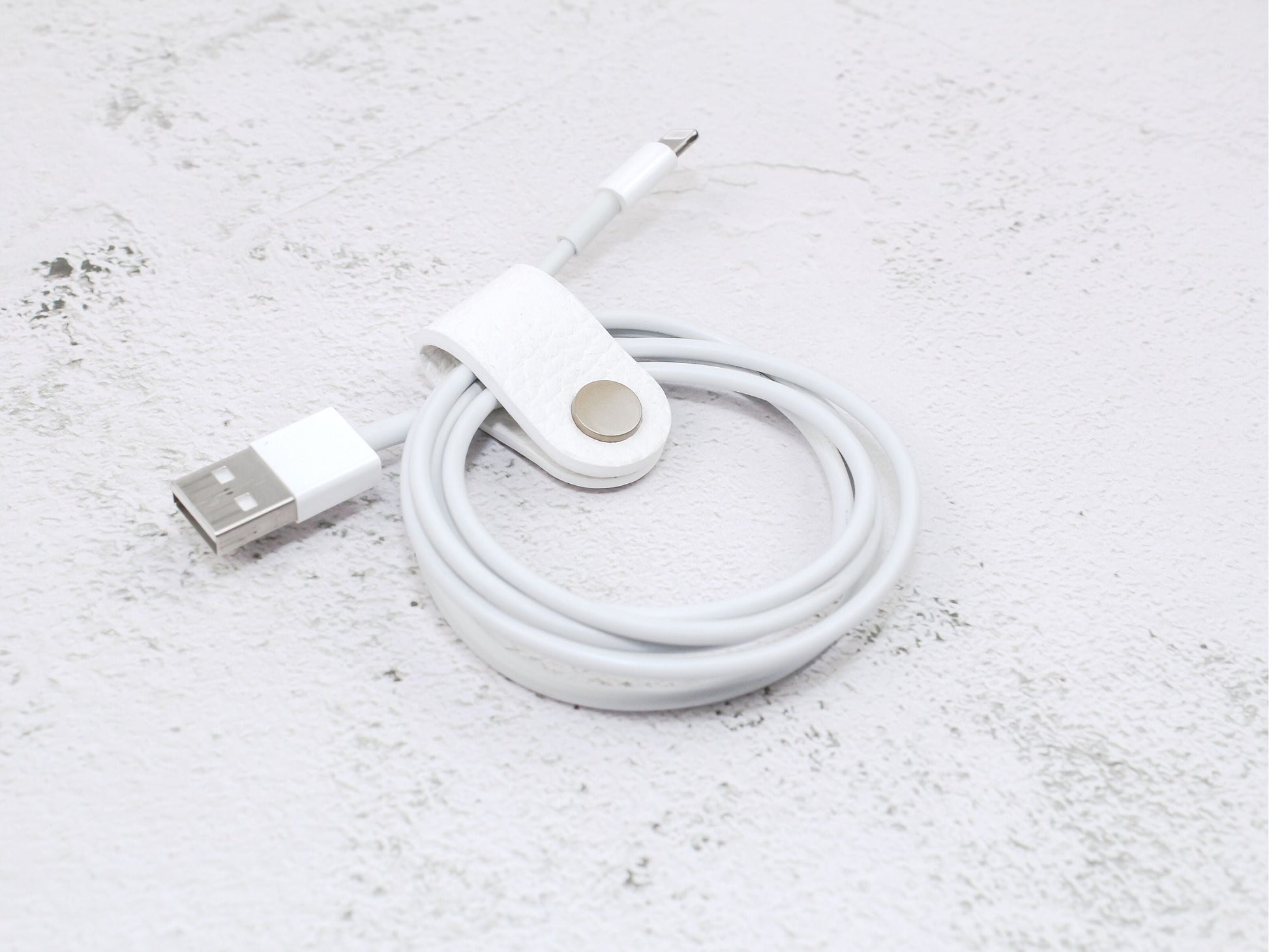 A white pebbled grain faux leather cord organiser wrap with a snap closure, designed to keep USB cables and earphone cords neat and tangle-free.